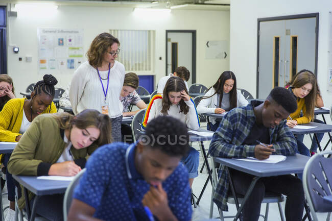 Profesora de secundaria supervisando estudiantes que toman exámenes en escritorios - foto de stock