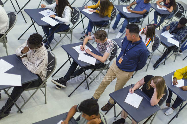 Profesora supervisando a estudiantes de secundaria que toman exámenes en escritorios - foto de stock