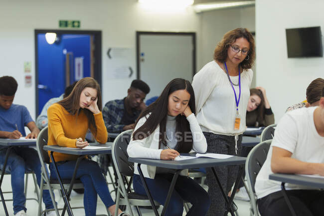 Professora do ensino médio supervisionando alunos fazendo exame em mesas — Fotografia de Stock