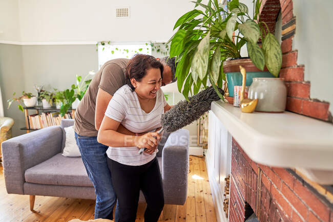 Affectionate playful mature couple with duster dusting living room — Stock Photo