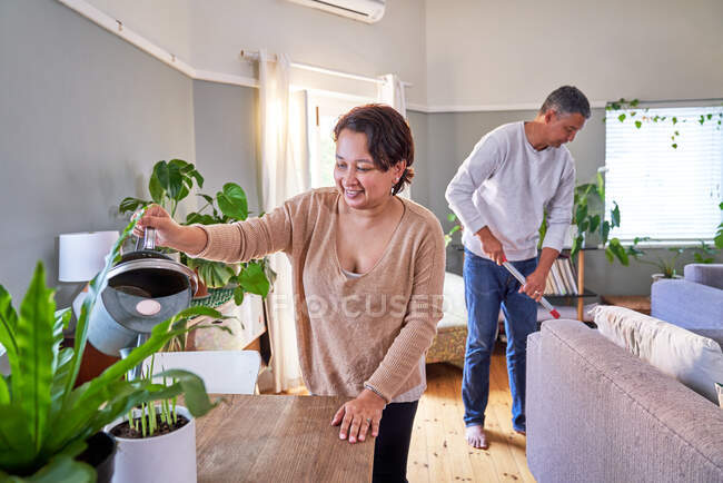 Couple d'âge mûr arrosage des plantes d'intérieur et de nettoyage salon — Photo de stock