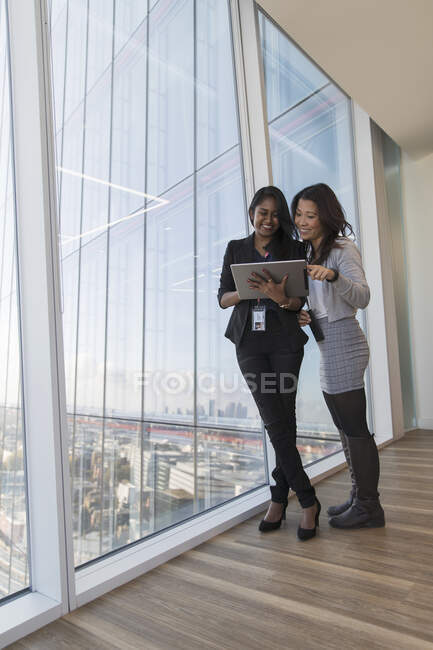 Mujeres de negocios sonrientes usando tableta digital en ventana de oficina de rascacielos - foto de stock