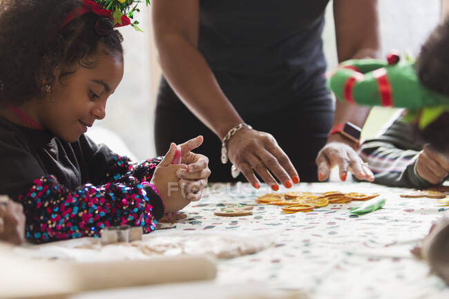 Chica decorando galletas de Navidad con la familia en la mesa - foto de stock