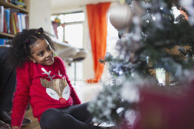 Neugieriges Mädchen im Weihnachtspullover blickt auf Weihnachtsbaum — Stockfoto