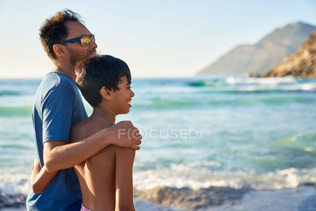 Padre e hijo cariñosos abrazándose en la soleada playa del océano - foto de stock