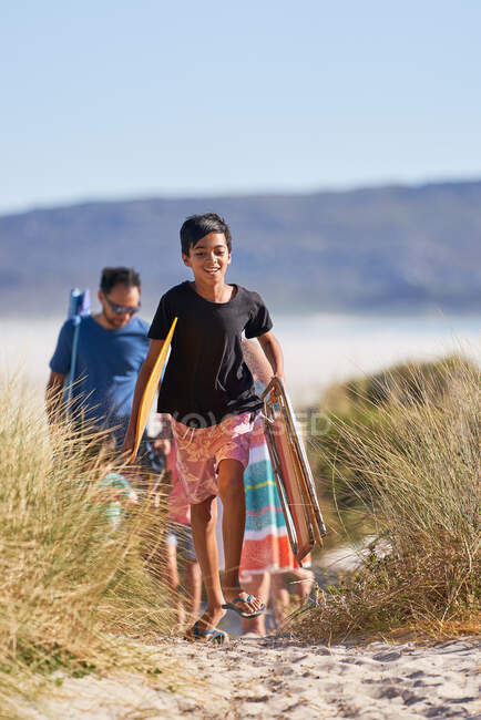 Menino feliz carregando cadeira dobrável e toalha na praia ensolarada — Fotografia de Stock