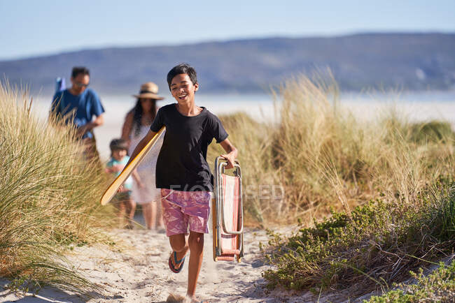 Счастливый мальчик бегает со складным стульчиком по солнечной пляжной дорожке — стоковое фото