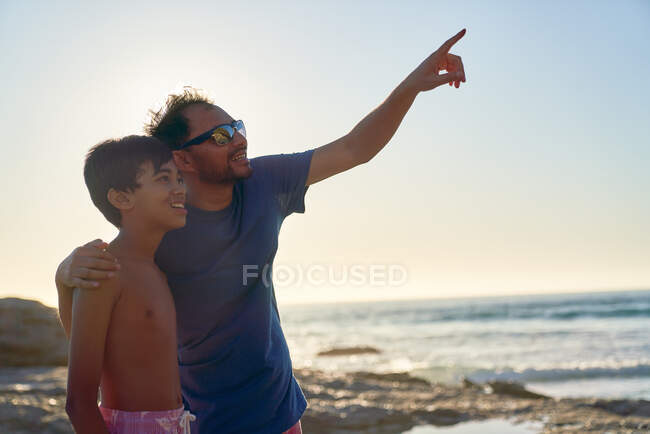 Curioso padre e hijo apuntando hacia el cielo en la soleada playa del océano - foto de stock