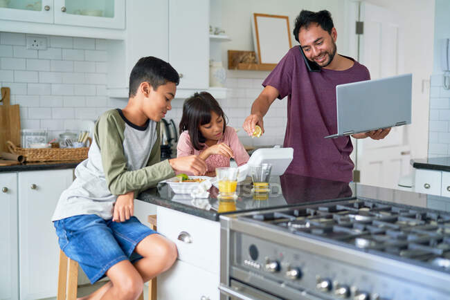 Pai com laptop alimentando crianças com tirar comida na cozinha — Fotografia de Stock