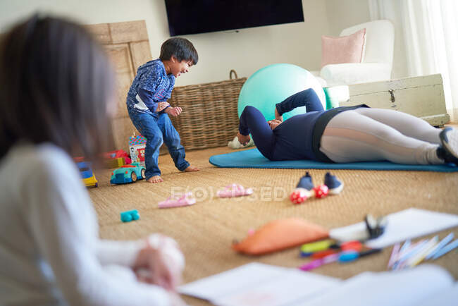 Verspielter Junge beobachtet Mutter beim Turnen auf Wohnzimmerboden — Stockfoto