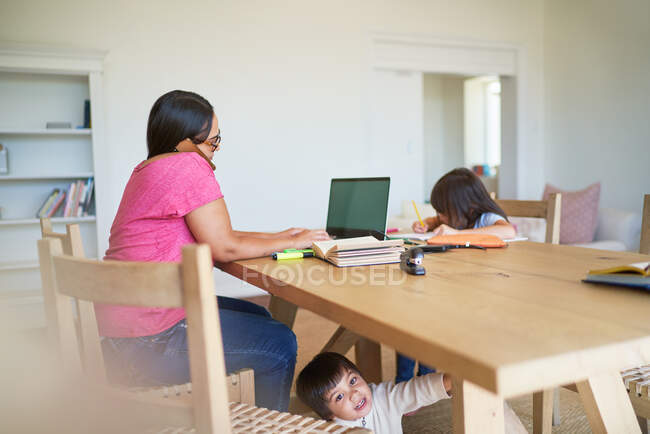 Mãe trabalhando no laptop com crianças brincando e fazendo lição de casa — Fotografia de Stock