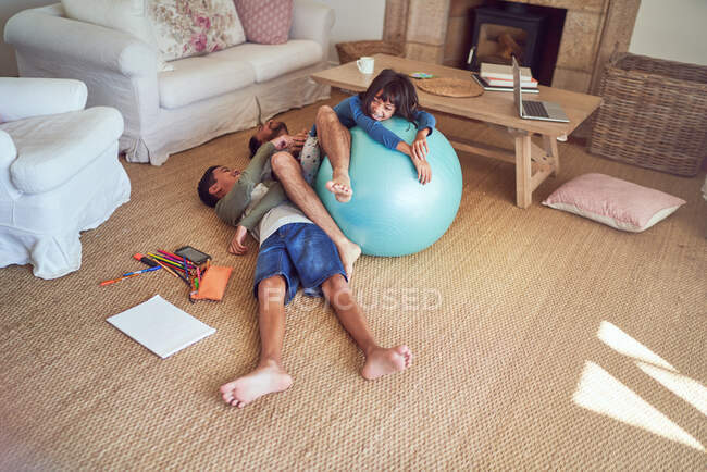 Padre juguetón y niños con pelota de fitness en el piso de la sala de estar - foto de stock