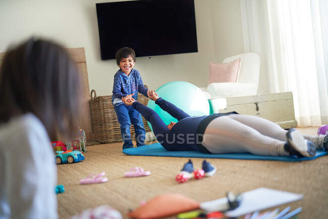 Ragazzo che gioca con la madre che si esercita sul tappetino yoga in soggiorno — Foto stock