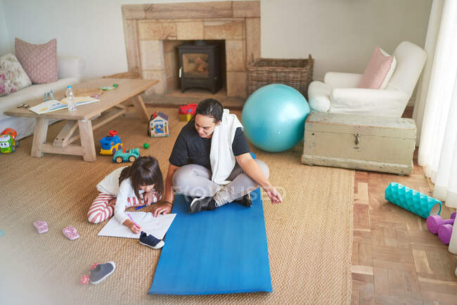 Madre su tappetino yoga guardando figlia colorare sul pavimento del soggiorno — Foto stock
