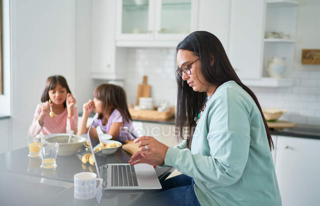 Madre trabajando en el ordenador portátil en la cocina mientras las hijas desayunan - foto de stock