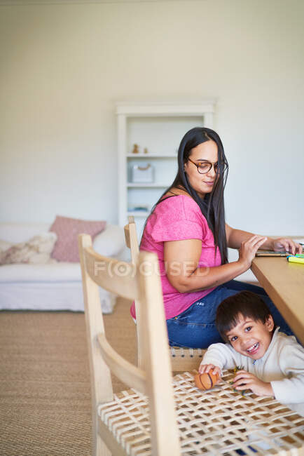 Hijo jugando debajo de la mesa de comedor mientras mamá trabaja - foto de stock
