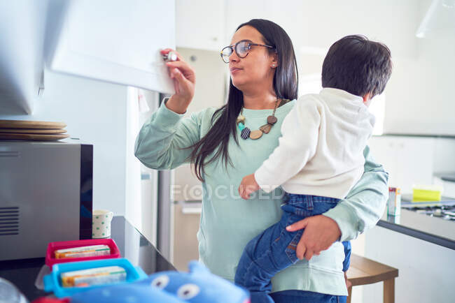 Mãe segurando filho e preparando almoços na cozinha — Fotografia de Stock