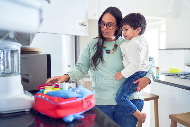 Mãe segurando filho e preparando o almoço escolar na cozinha — Fotografia de Stock