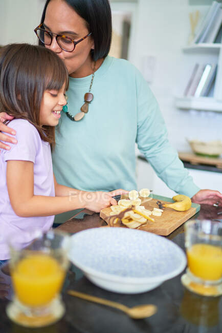 Прихильна мати цілує дочку, що ріже банан на кухні — стокове фото