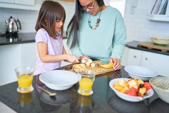 Mother helping daughter cut banana in kitchen — Stock Photo