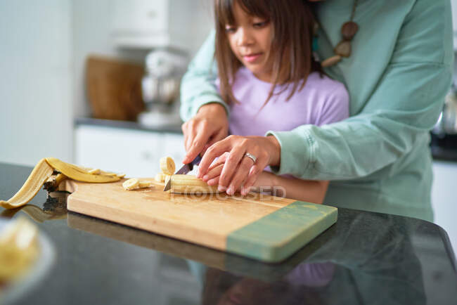 Mãe ajudando filha cortar banana na cozinha — Fotografia de Stock