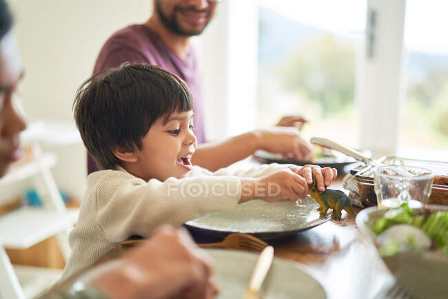 Счастливый мальчик играет с игрушечным динозавром за обеденным столом — стоковое фото
