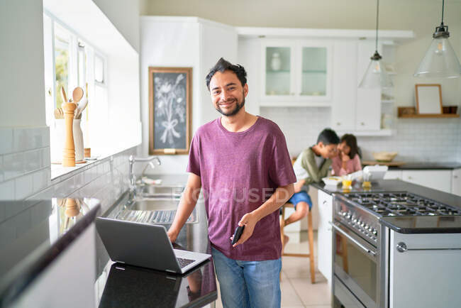 Retrato feliz padre trabajando en el ordenador portátil en la cocina con los niños comiendo - foto de stock