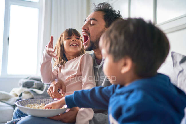 Niños juguetones alimentando palomitas de maíz padre en el sofá - foto de stock