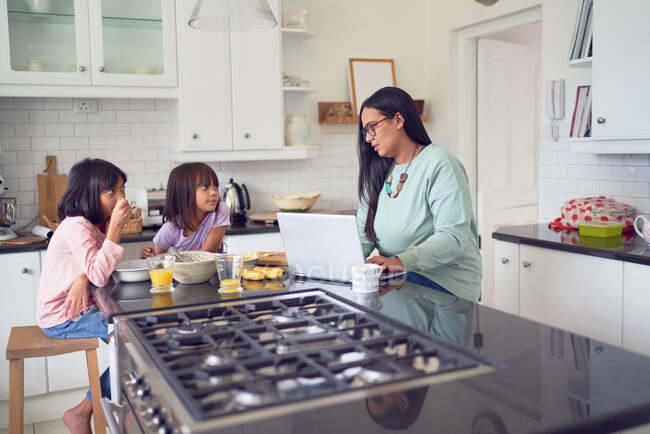 Mãe trabalhando no laptop, enquanto as filhas tomam café da manhã na cozinha — Fotografia de Stock