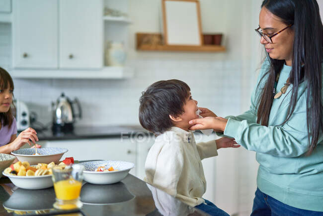 Madre che asciuga la faccia di figlio che mangia frutta in cucina — Foto stock