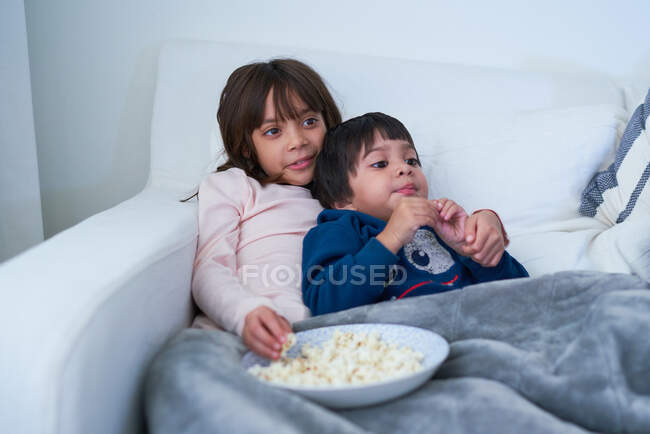 Cariñoso hermano y hermana comiendo palomitas de maíz y viendo la televisión en el sofá - foto de stock