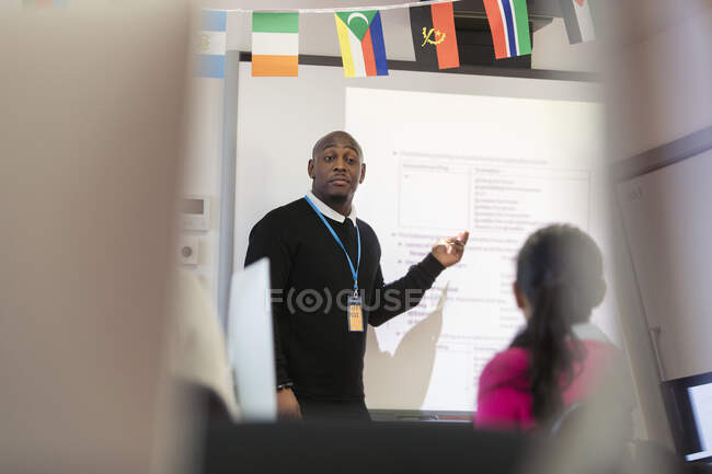 Instructor de universidad comunitaria masculino que lidera la lección en la pantalla de proyección en el aula - foto de stock