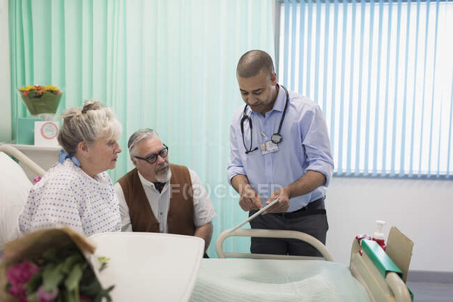 Doctor con tableta digital haciendo rondas, hablando con pareja mayor en la habitación del hospital - foto de stock