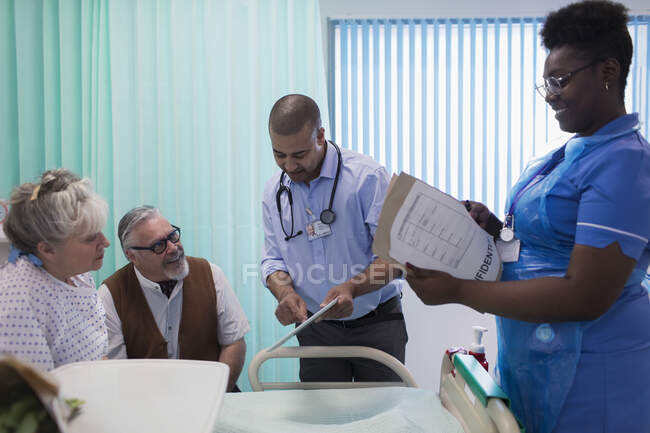 Médico y enfermera con tableta digital y historial médico haciendo rondas, hablando con pareja mayor en la habitación del hospital - foto de stock