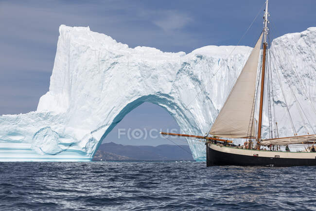 Корабль проплывает мимо солнечной величественной айсбергской арки на океане Гренландии — стоковое фото