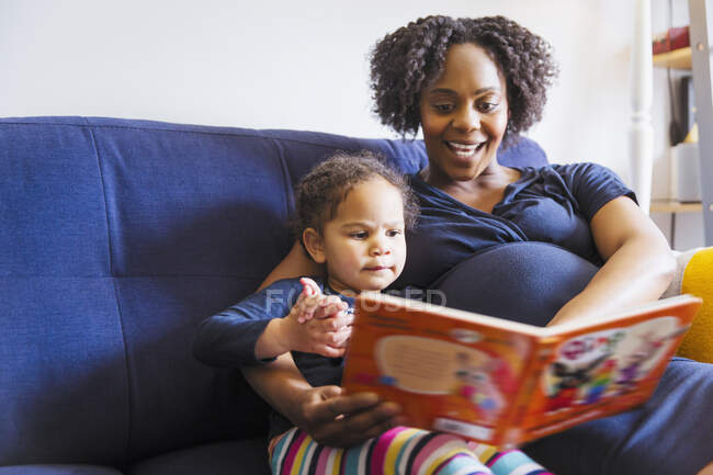 Embarazada madre leyendo libro a hija en sofá - foto de stock