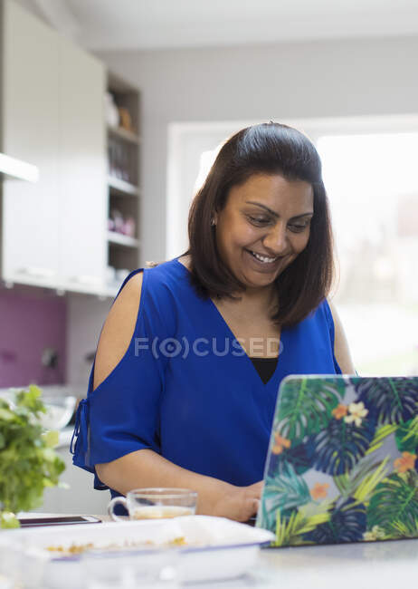Усміхнена жінка використовує ноутбук на кухні — стокове фото