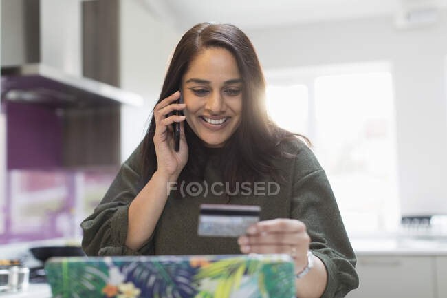 Mulher sorridente com cartão de crédito e telefone inteligente pagando contas na cozinha — Fotografia de Stock