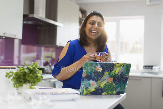 Happy woman with tea laughing at laptop in kitchen — Stock Photo