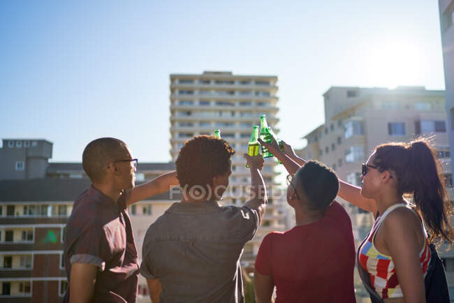 Jóvenes amigos brindando botellas de cerveza en el soleado balcón de la azotea urbana - foto de stock