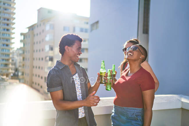 Jovens amigos felizes bebendo cerveja na varanda ensolarada do telhado urbano — Fotografia de Stock