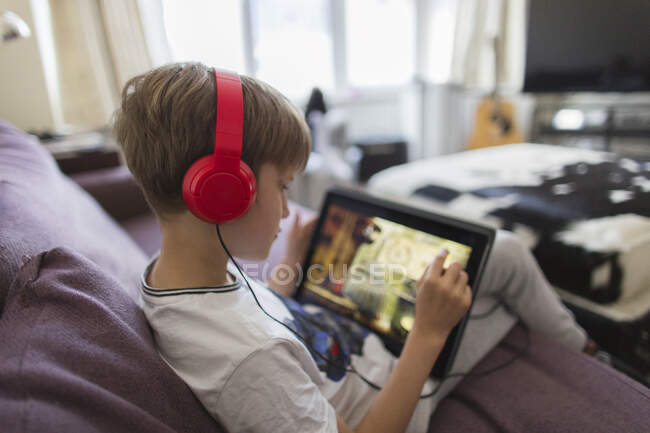 Boy with headphones and digital tablet playing video game on sofa — Stock Photo