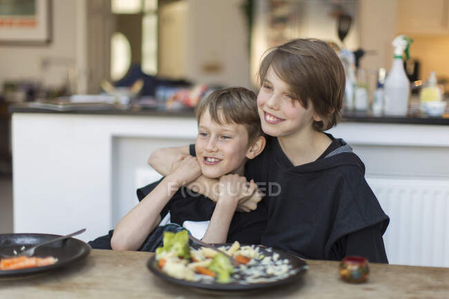 Happy brothers hugging and eating dinner at dining table — Stock Photo