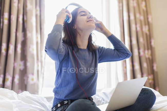 Menina adolescente sorridente com fones de ouvido e laptop na cama ensolarada — Fotografia de Stock