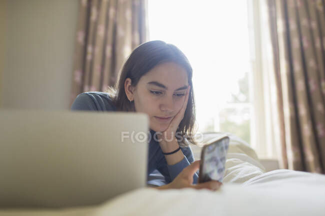 Девочка-подросток с помощью смартфона на ноутбуке на кровати — стоковое фото