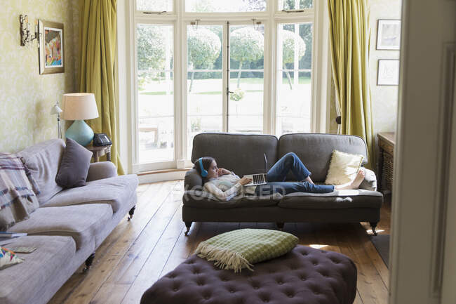 Adolescente avec écouteurs et ordinateur portable relaxant sur le canapé du salon — Photo de stock