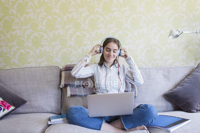 Menina adolescente sorridente com fones de ouvido usando laptop no sofá da sala de estar — Fotografia de Stock