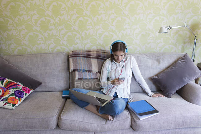 Chica adolescente con auriculares y portátil con teléfono inteligente en el sofá - foto de stock