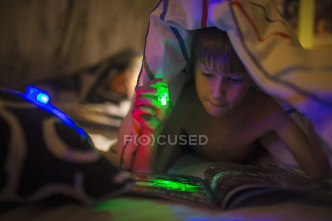Menino com lanterna verde leitura revista sob cobertor — Fotografia de Stock