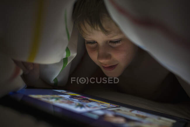 Закрыть мальчика цифровой табличкой под одеялом — стоковое фото
